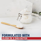 A clear glass clamp-lid jar filled with white scrub or balm, shown on a light marble-like surface with a wooden spoon holding some of the product. Text at the bottom reads 'FORMULATED WITH JOJOBA OIL & SHEA BUTTER'.