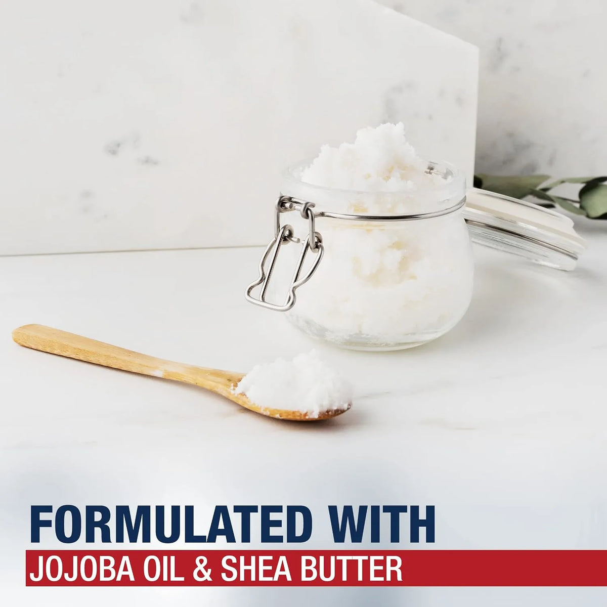 A clear glass clamp-lid jar filled with white scrub or balm, shown on a light marble-like surface with a wooden spoon holding some of the product. Text at the bottom reads 'FORMULATED WITH JOJOBA OIL & SHEA BUTTER'.